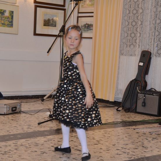 Ein kleines Mädchen in einem sternförmigen Kleid steht auf der Bühne, neben einem Mikrofon und einer Gitarre. Hinter ihr hängen gerahmte Bilder an einer weißen Wand.