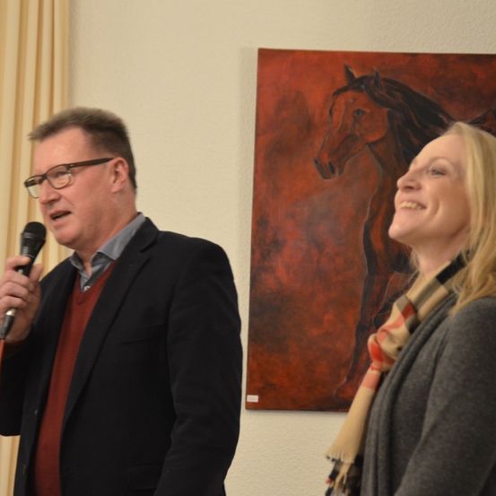 Ein Mann in einem schwarzen Anzug spricht in ein Mikrofon, neben ihm steht eine Frau in einem grauen Cardigan und einem Schal. Ein rotes Pferdebild hängt an der Wand hinter ihnen.