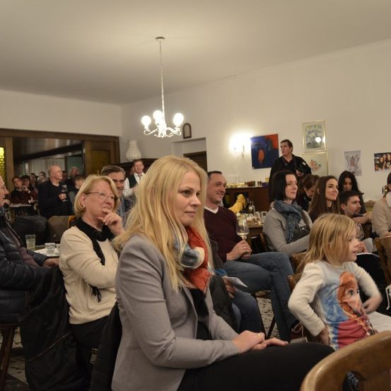 Eine Zusammenkunft in Innenräumen mit Menschen, die auf Stühlen sitzen, eine Frau mit blonden Haaren vorne, ein junges Mädchen mit einem Disney-Charakter-Shirt, einer von der Decke hängenden Kronleuchter und mehreren Bildern an der Wand.