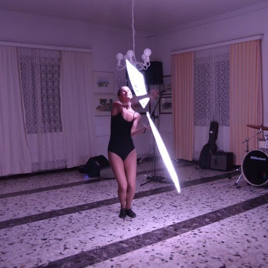 Eine Frau in einem schwarzen Badeanzug posiert in einem Raum mit einem Lichtschwert und einem Lautsprecher. Im Hintergrund befinden sich ein Schlagzeug und eine Gitarrenkoffer.