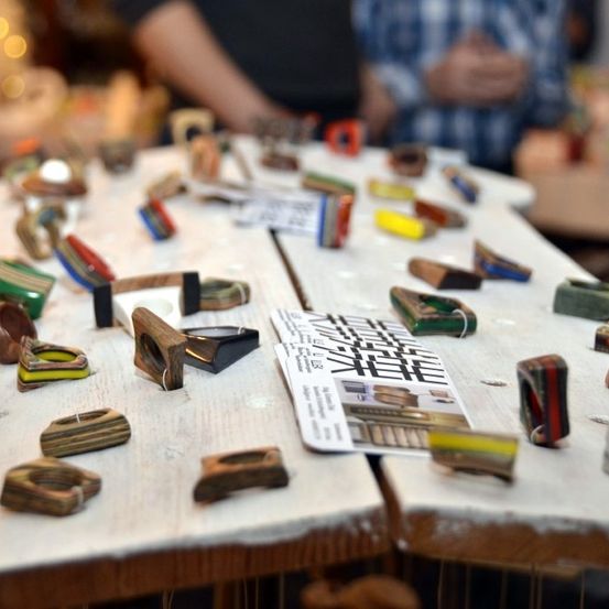 Ein Tisch zeigt verschiedene Holz- und Harzobjekte mit unterschiedlichen Farben und Designs. Ein Stück Papier mit Text und Bildern ist ebenfalls vorhanden. Zwei Personen stehen hinter dem Tisch.
