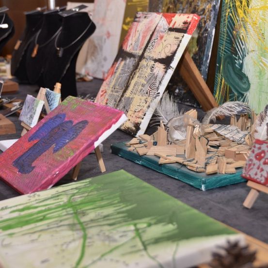 Eine Ausstellung verschiedener Kunstwerke, darunter farbenfrohe Gemälde und eine Mixed-Media-Skulptur, auf einem Tisch mit Holzstaffeln und Halsketten im Hintergrund angeordnet.