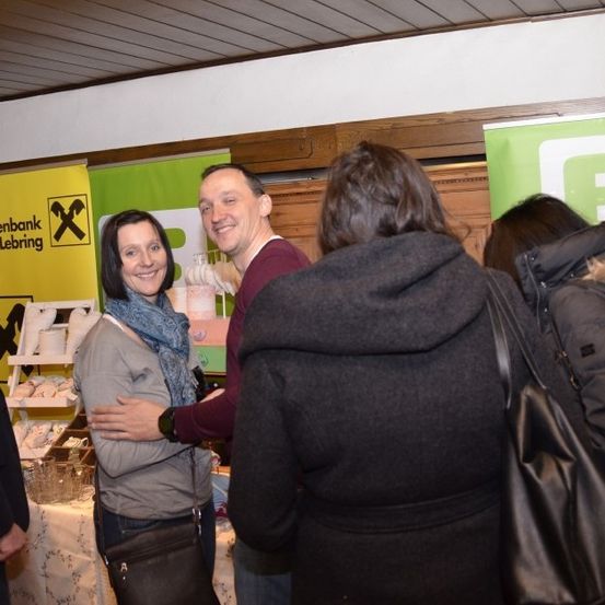 Mehrere Menschen stehen in einem Raum, vielleicht bei einer Veranstaltung. Eine Frau lächelt und steht in der Nähe eines Tisches mit mehreren Gegenständen darauf. Ein Mann in einem bordeauxfarbenen Hemd umarmt eine andere Frau.