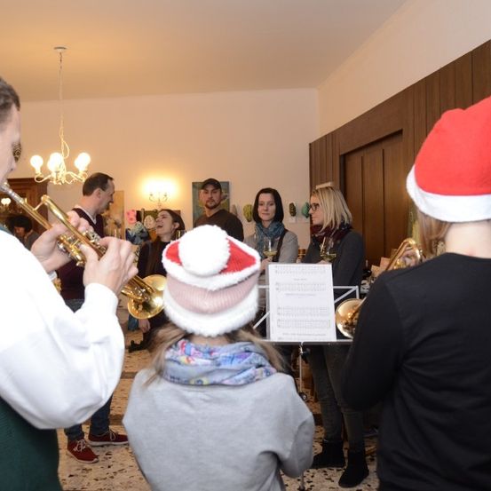 Eine Gruppe von Menschen versammelt sich drinnen, einige tragen festliche Mützen und lauschen einem Trompeter, während andere Posaunen und Trompeten spielen.