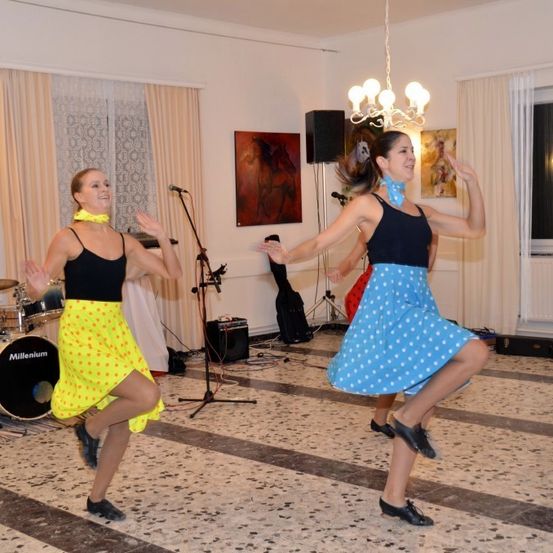 Zwei Frauen tanzen in einem Raum mit einem Schlagzeug und Mikrofon, während eine weitere Frau hinter ihnen tanzt. Eine Lampe und Kunstwerke schmücken die Wände.