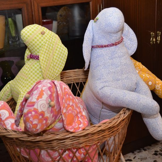 Ein Korb aus Weideholz enthält drei gehäkelte Hasen, jeweils mit unterschiedlichen Mustern und Farben, vor einem Hintergrund aus einem Holzschrank.