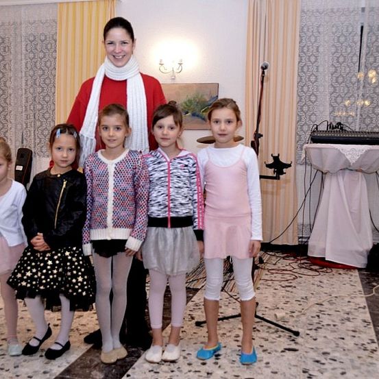 Eine Gruppe junger Mädchen in verschiedenen Outfits steht neben einer Frau in einem roten Pullover in einem Raum mit einer musikalischen Einrichtung im Hintergrund.