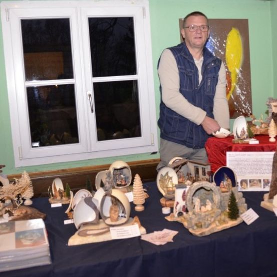 Ein Mann steht hinter einem mit blauem Tuch bedeckten Tisch. Der Tisch ist mit verschiedenen Skulpturen und handgefertigten Gegenständen gefüllt, möglicherweise zum Verkauf. Der Raum hat grüne Wände und ein Fenster mit weißen Rahmen.