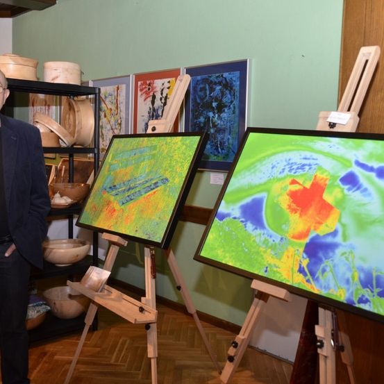 Ein Mann in Anzug steht neben einer Ausstellung von Gemälden, hält ein Weinglas und steht in einem Raum mit Holzböden.