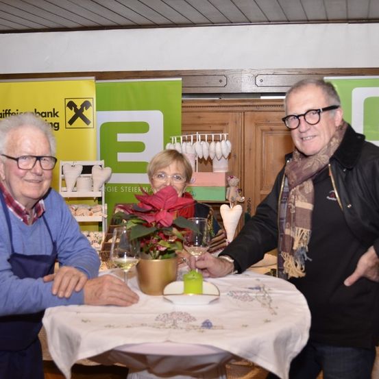 Drei Menschen stehen um einen Tisch mit einer Blume, Gläsern und einem Teller. Der Mann rechts trägt eine Krawatte und eine Brille. Die Frau hinter dem Tisch trägt eine Brille. Ein gelber und grüner Banner steht hinter ihnen.