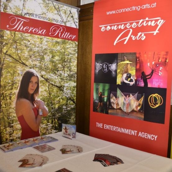 Eine Werbetafel für Theresa Ritter und Connecting Arts zeigt eine Frau in Rot, einen Tisch mit Broschüren und einen Hintergrund aus Bäumen.