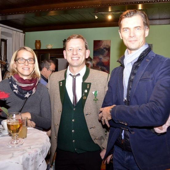 Drei Menschen stehen nebeneinander lächelnd in einem Raum mit einem Tisch mit Getränken und Blumen. Der Mann in der Mitte trägt ein grünes Weste mit einer Krawatte und Knöpfen. Hinter ihnen ist eine grüne Wand mit einem Regal und einer Vase.