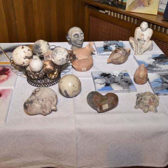 Ein Tisch, der mit einem weißen Tuch bedeckt ist, zeigt verschiedene Keramikskulpturen, darunter ein Korb mit runden Objekten, ein herzförmiges Stück und eine Schildkröte. Auf dem Tisch befinden sich auch abstrakte Gemälde und ein Regal mit weiteren Kunstwerken im Hintergrund.
