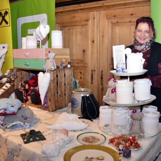 Eine Frau steht hinter einem Tisch voller verschiedener handgefertigter Artikel, darunter weiße Keramikgläser, Teller, Plüschtiere und eine Kerze. Dahinter steht ein hölzerner Schrank mit einem Banner und einem Holzregal mit Gegenständen.