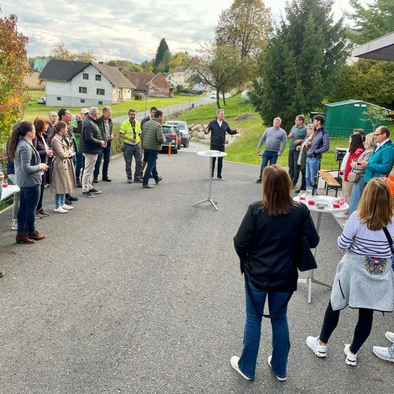 Eine Gruppe von Menschen versammelt sich auf einem Parkplatz, wobei ein Mann vor ihnen spricht. Sie halten Getränke und unterhalten sich, während andere hinter Tischen stehen. Bäume und Häuser sind im Hintergrund zu sehen.