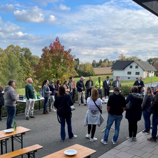 Eine Gruppe von Menschen steht und unterhält sich im Freien. Es gibt Tische mit Tellern und Flaschen. Dahinter steht ein Haus und ein geparktes Auto. Der Himmel ist blau mit Wolken.