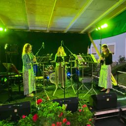 Drei Frauen stehen mit Instrumenten und einem Schlagzeug in einer grün beleuchteten Umgebung auf der Bühne. Eine spielt Klarinette, eine andere Flöte, und die dritte ist rechts. Musiknoten stehen auf Ständern und Lautsprecher sind auf der Bühne.