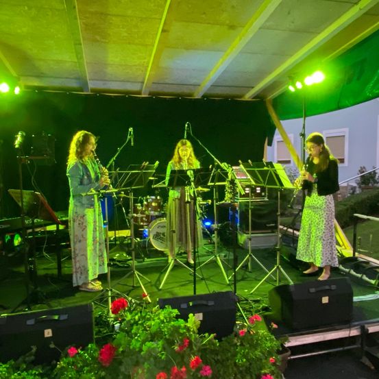 Drei Frauen stehen mit Instrumenten und einem Schlagzeug in einer grün beleuchteten Umgebung auf der Bühne. Eine spielt Klarinette, eine andere Flöte, und die dritte ist rechts. Musiknoten stehen auf Ständern und Lautsprecher sind auf der Bühne.