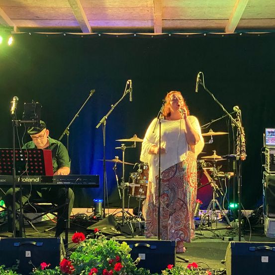 Eine Band tritt auf der Bühne auf, eine Frau singt in ein Mikrofon. Ein Mann spielt vor sich das Keyboard. Verschiedene Musikinstrumente und Ausrüstungsgegenstände befinden sich auf der Bühne.