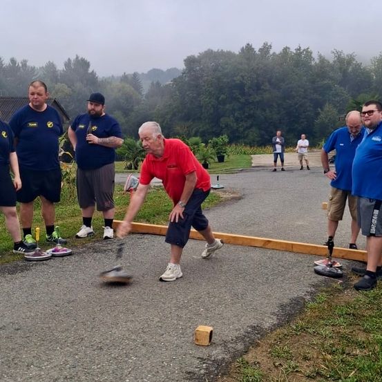 Ein älterer Mann wirft einen Kugelstoß, während andere zusehen, im Freien. Bäume und Gras sind im Hintergrund.