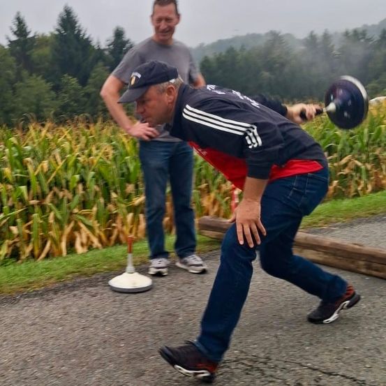 Ein Mann beugt sich vor und wirft ein Gewicht in einem Maisfeld. Ein zweiter Mann steht hinter ihm und lächelt.