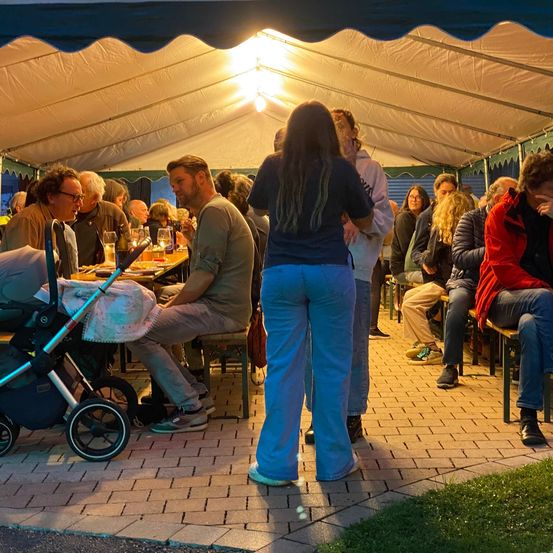 Unter einem Zelt versammelt sich eine Gruppe von Menschen zum Abendessen. Einige sitzen, andere stehen, und ein Kinderwagen ist in der Nähe. Ein Mann mit Brille sitzt am Tisch.