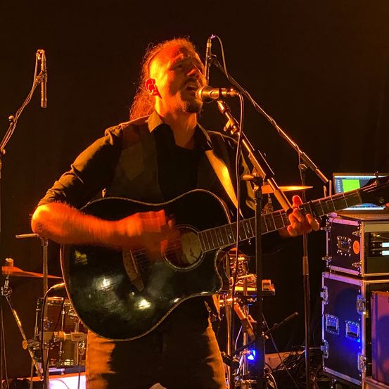 Ein Musiker mit langen Haaren und Bart steht auf der Bühne und spielt Gitarre. Er singt in ein Mikrofon. Dahinter sind Trommeln, ein Monitor und weitere Musikgeräte. Die Bühne ist schwach beleuchtet.