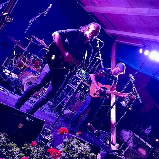 Eine Band tritt auf der Bühne bei lila Beleuchtung auf, ein Mann spielt Gitarre und singt in ein Mikrofon. Ein anderer Mann spielt Gitarre dahinter.