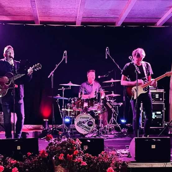 Eine Band spielt auf einer Bühne unter lila Beleuchtung. Einer spielt Gitarre, ein anderer Schlagzeug und der dritte Bassgitarre. Mikrofone und Lautsprecher sind vorhanden.