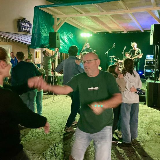 Eine Gruppe von Menschen tanzt bei Nacht draußen, mit einer Band, die auf der Bühne im Hintergrund auftritt. Die Bühne ist mit einem grünen Tarp bedeckt und hat Lautsprecher und ein Mikrofon-Setup.
