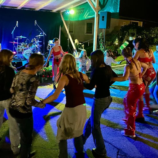 Eine Nachtveranstaltung mit einer Band auf der Bühne. Mehrere Frauen in verschiedenen Outfits tanzen vor der Bühne. Ein Lautsprecher steht in der Nähe der Bühne.