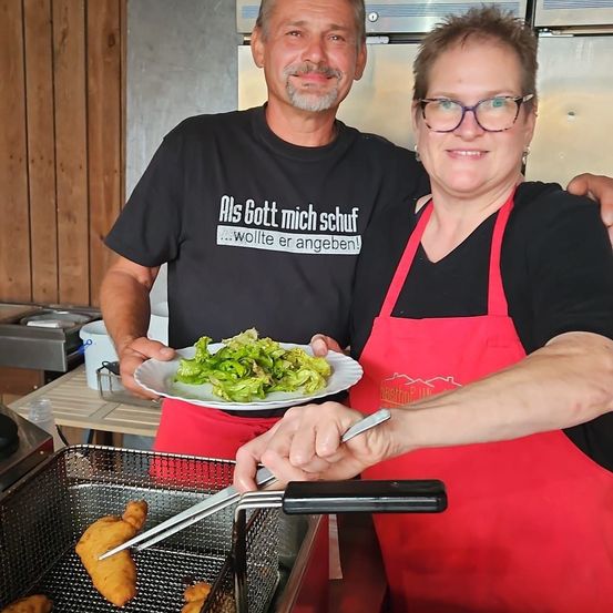 Ein Mann und eine Frau in Schürzen stehen hinter einer Frittierstation mit einem Teller grünem Salat und frittierten Lebensmitteln in einer Küche.
