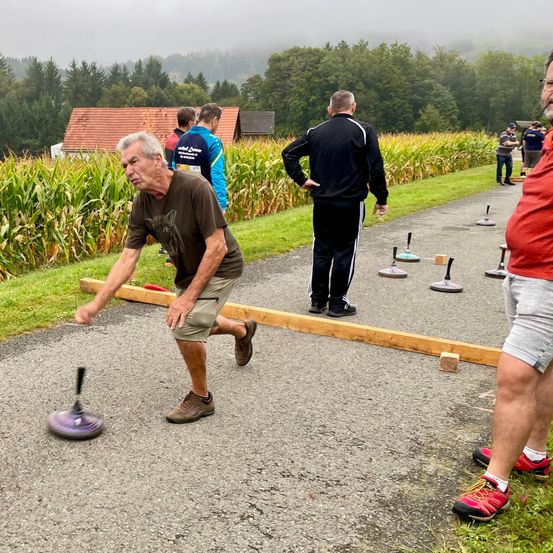 Ein älterer Mann wirft einen Bocce-Ball in einem Spiel, das in einem Maisfeld aufgebaut ist. Hinter ihm beobachten oder nehmen andere teil.