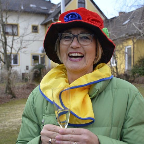 Eine Frau in einem roten Hut und einer grünen Jacke mit einem gelben Schal steht draußen, hält ein Weinglas in der Hand und lächelt mit einem Haus im Hintergrund.