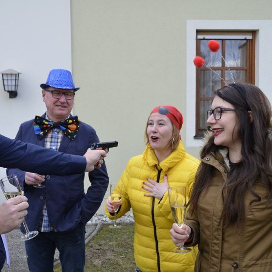 Vier Personen stehen draußen, eine hält eine Pistole und zielt auf einen Mann mit einer Schleife, während zwei Frauen lachen und Champagner-Gläser halten.