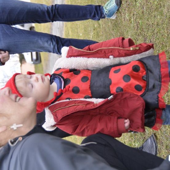 Ein Kleinkind in einem roten Marienkäfer-Kostüm mit schwarzen Punkten liegt auf Gras, neben einer Frau mit Kreuz-Halskette.