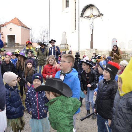 Eine Gruppe von Kindern in verschiedenen Kostümen versammelt sich vor einer Kirche mit einem Kreuz und einer Statue.