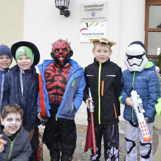 Eine Gruppe von Kindern steht vor einem Gebäude. Einige von ihnen tragen Masken und halten Spielzeug. Über ihnen befindet sich ein Schild.