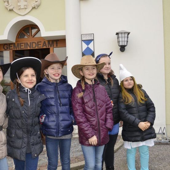 Sechs junge Mädchen stehen vor dem Gemeindeamt-Gebäude, einige tragen Cowboyhüte und andere weiße Partyhüte. Das Mädchen in der Mitte trägt eine lila Pufferjacke.
