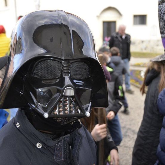 Eine Person mit einer Darth-Vader-Maske steht in einer Menschenmenge im Freien. Im Hintergrund befinden sich ein Gebäude und mehrere Personen.