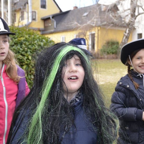 Drei junge Mädchen in Kostümen, eines mit grünen Haaren, stehen vor einem gelben Haus.