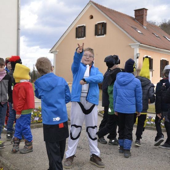Mehrere Kinder in Winterkleidung stehen vor einem Haus, wobei ein Junge ein blaues Kostüm trägt und ein Friedenszeichen macht.