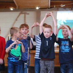 Mehrere Kinder stehen in einer Reihe, wobei eines von ihnen ein schwarzes T-Shirt mit der Nummer 1 trägt. Sie machen eine Herzform mit ihren Händen. Dahinter steht ein Holztisch mit einem Mikrofon.