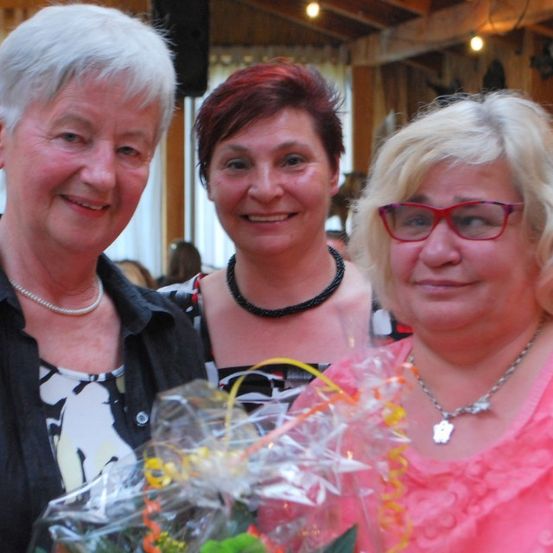 Drei lächelnde Frauen vor der Kamera, eine hält einen Blumenstrauß, eine andere mit roten Haaren, alle tragen Halsketten.