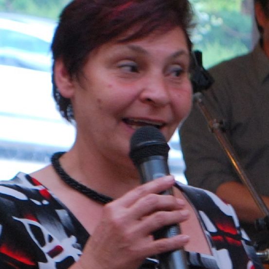 Eine Frau mit kurzen roten Haaren hält ein Mikrofon. Sie lächelt und scheint zu sprechen. Hinter ihr hält eine Person in einem braunen Hemd eine Posaune.