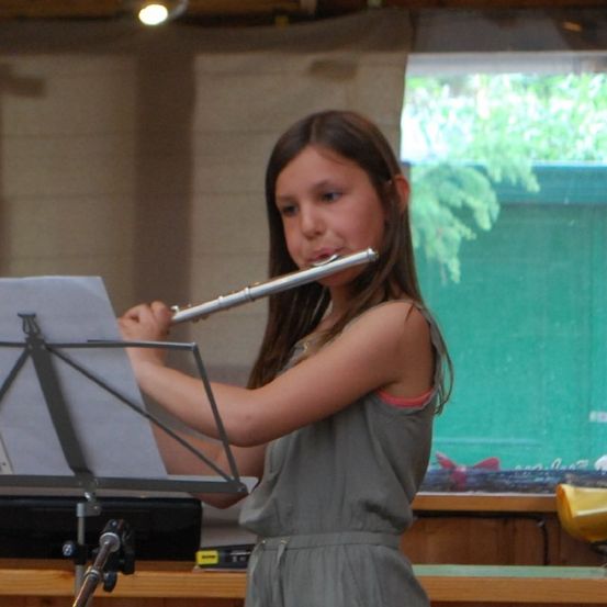 Ein Mädchen steht in einem Raum und spielt eine Flöte, während es Notenblätter betrachtet. Der Raum hat einen Holztisch und ein Fenster mit grüner Aussicht.