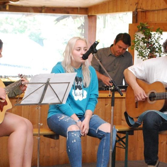 Vier Personen sitzen auf einer Holzbank und spielen Gitarre. Eine Frau singt in ein Mikrofon. Eine weitere Frau und zwei Männer spielen Gitarre. Ein Topf mit Pflanzen und ein Mikrofonständer stehen hinter ihnen.