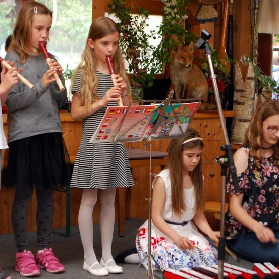 Fünf Mädchen stehen in einem Raum und spielen jeweils eine Blockflöte. Ein Mädchen sitzt auf dem Boden und spielt ein Xylophon. Dahinter steht ein Tisch mit einem stofftier Fuchs und einem Mikrofon auf einem Ständer.