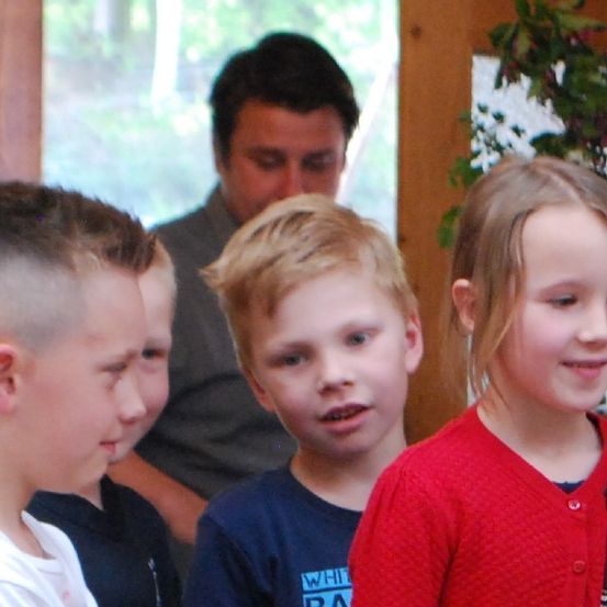 Vier Kinder in einem Raum, eines mit einem Mikrofon, ein Mann dahinter und eine Pflanze im Hintergrund.