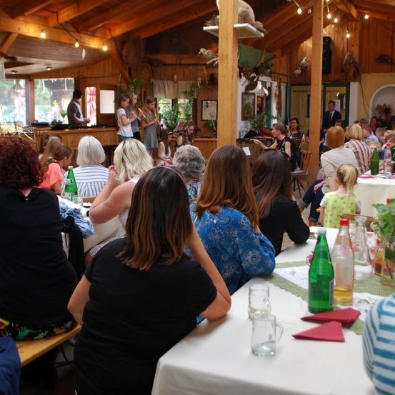 Eine Indoor-Veranstaltung mit einer Gruppe von Menschen, die an Tischen sitzen, möglicherweise in einem Restaurant, mit einer Person, die in der Mitte ein Musikinstrument spielt.